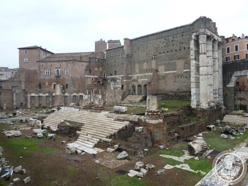 Ruined temple podium with a wide staircase and three standing columns against a high ancient wall of large stone blocks, modern buildings behind