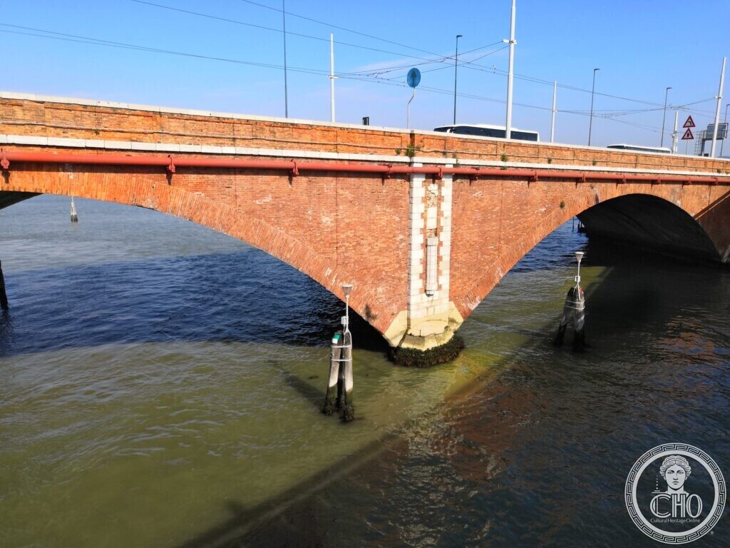 Two red brick arches of the bridge spanning green lagoon water, with a pale stone-faced pier and channel markers