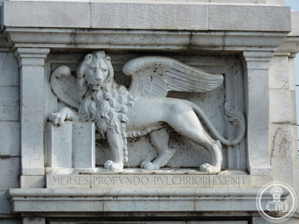 A white stone relief of the winged lion of Saint Mark with its paw on a book, above a band carved with a Latin motto