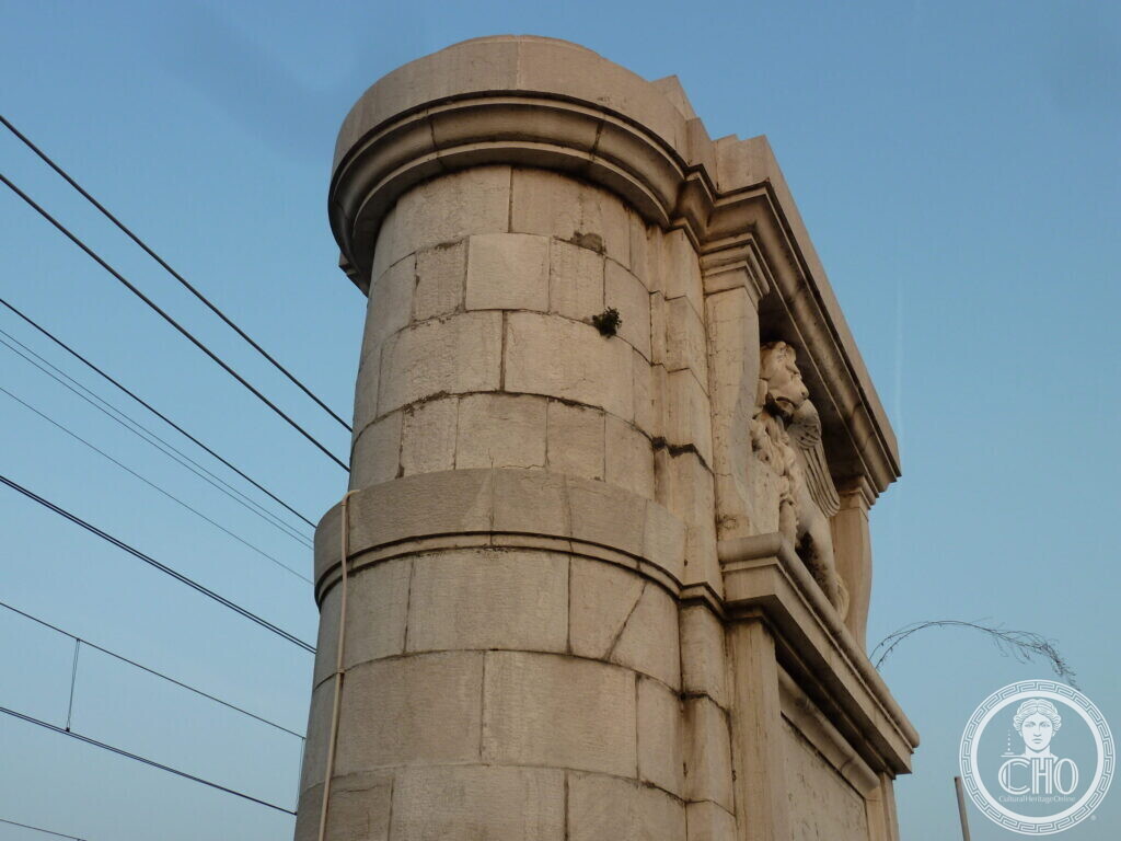 A tall rounded stone pylon of the Ponte della Liberta seen from below against a blue sky, crossed by overhead electric wires