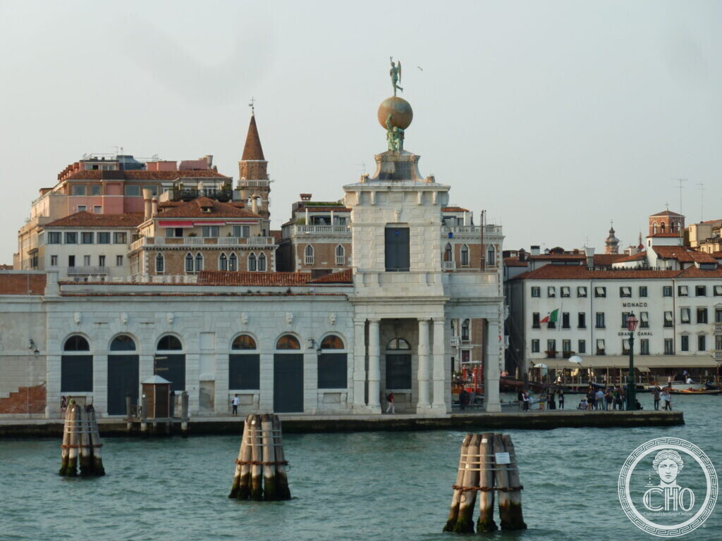 The white Dogana building with its globe tower in front of a row of Venetian palaces, mooring posts in the water