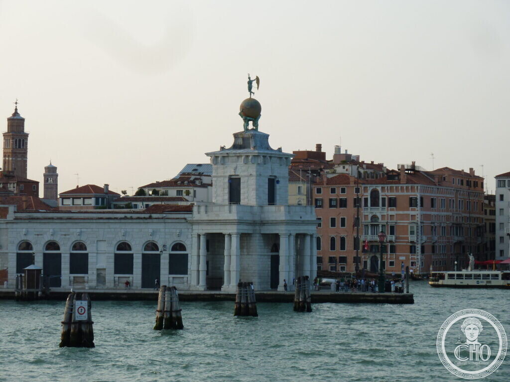 The Punta della Dogana seen across the water, a low white arcaded building with a tower and globe, wooden mooring posts in front