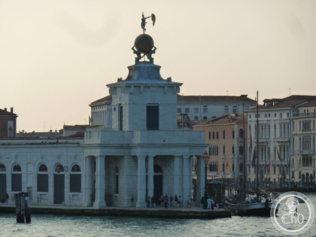 The white stone tower of the Punta della Dogana with a golden globe held by two bronze figures and a statue on top, people on the quay