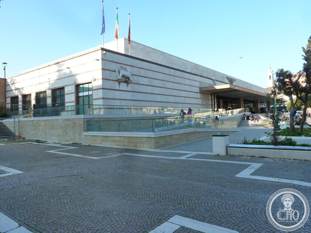 A low white stone building with horizontal bands and a relief of a winged lion, flags on the roof, a paved square in front