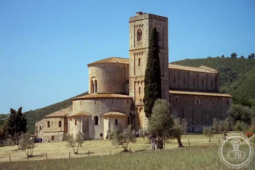 Abbazia di Sant Antimo, apse romanica in travertino sul fondovalle del Monte Amiata con cipresso e cielo toscano, Montalcino