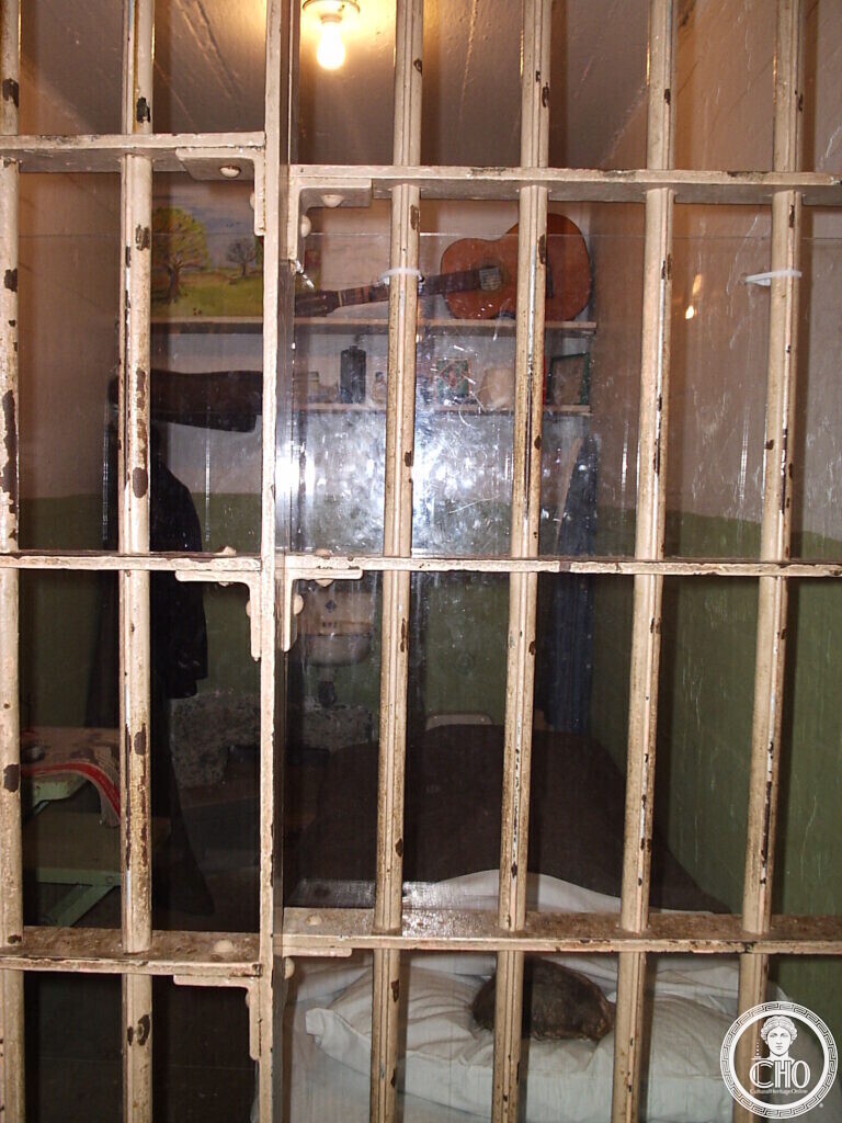 Prison cell seen through bars, with a bunk, a guitar hanging on the wall and small paintings