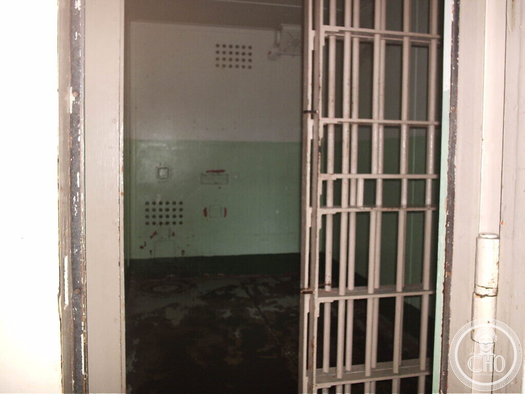 Small prison cell seen through an open barred door, pale green walls, a bunk and a ventilation grille