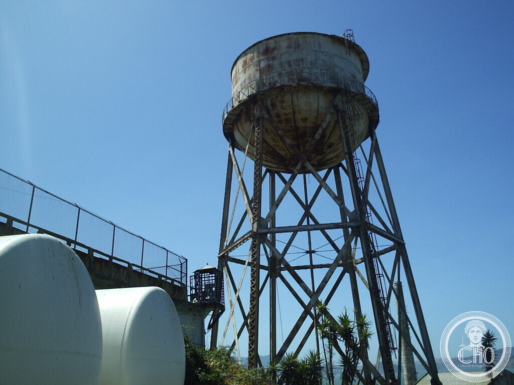 Rusting steel water tower on a lattice frame against a blue sky, with white tanks and a railing below