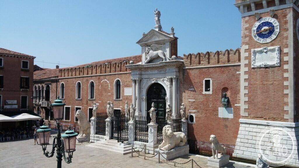 A white stone Renaissance gateway with a winged lion in a crenellated brick wall, marble lions in front, a brick tower with a blue clock on the right