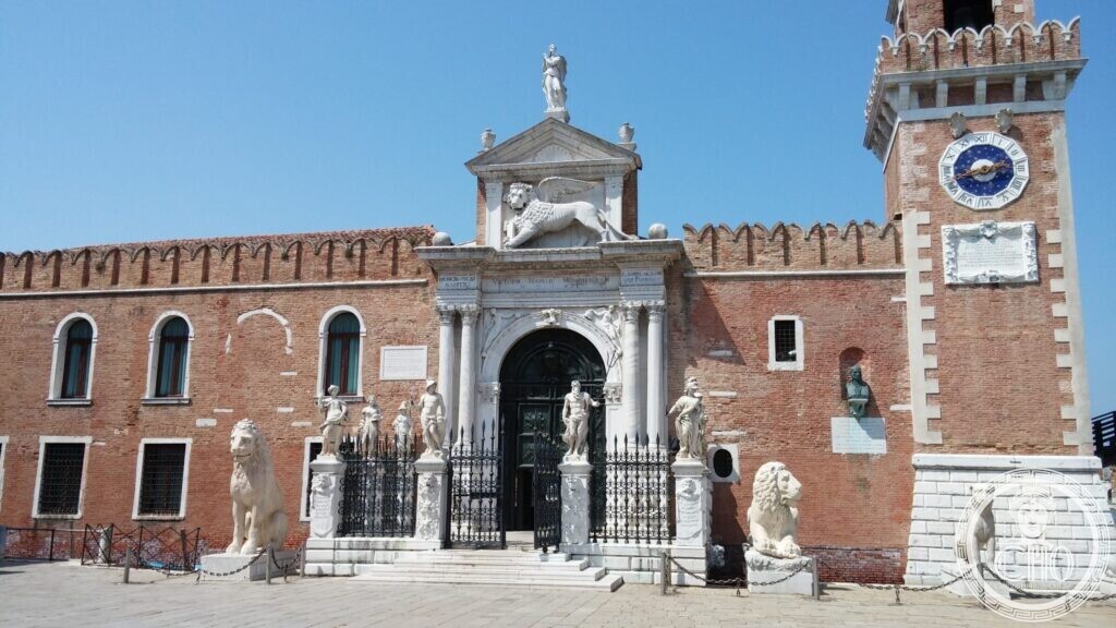 Frontal view of the white stone arched gate of the Venice Arsenal with statues on its railing, a winged lion above and lions on both sides