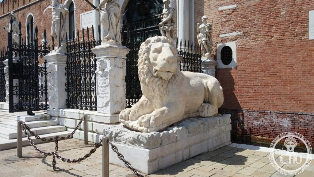 A large weathered white marble lion lying on a pedestal in front of the iron railing of the Arsenal gate