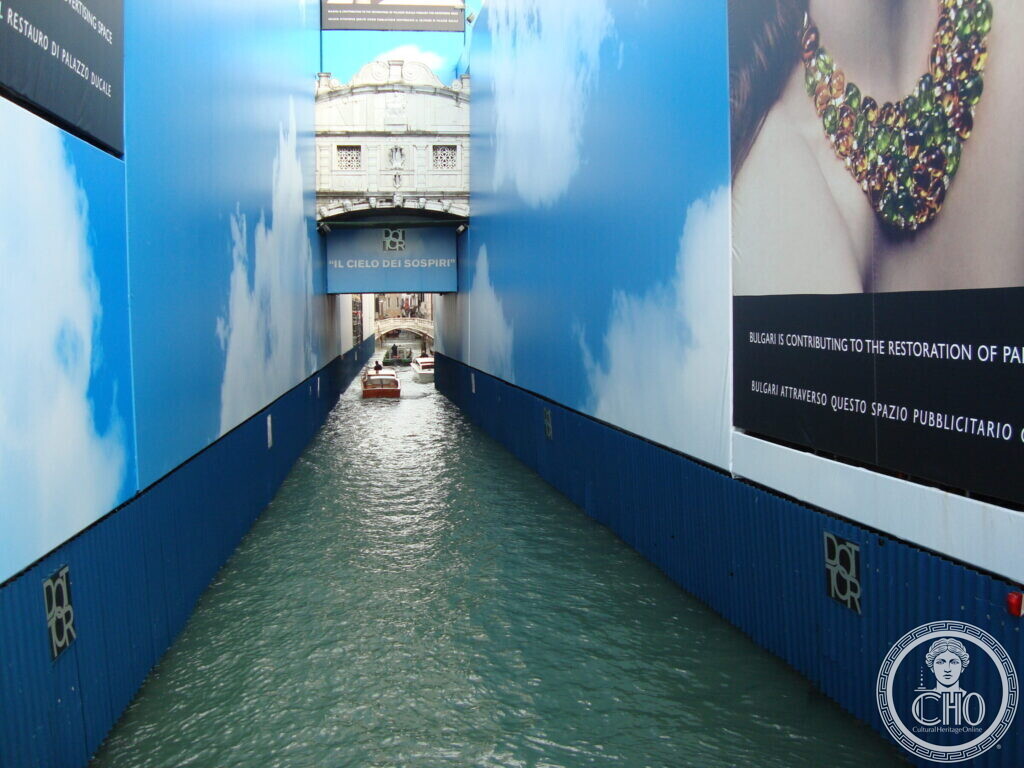 The Bridge of Sighs seen between two tall advertising hoardings printed with a blue sky and clouds that cover the scaffolding on either side of the canal