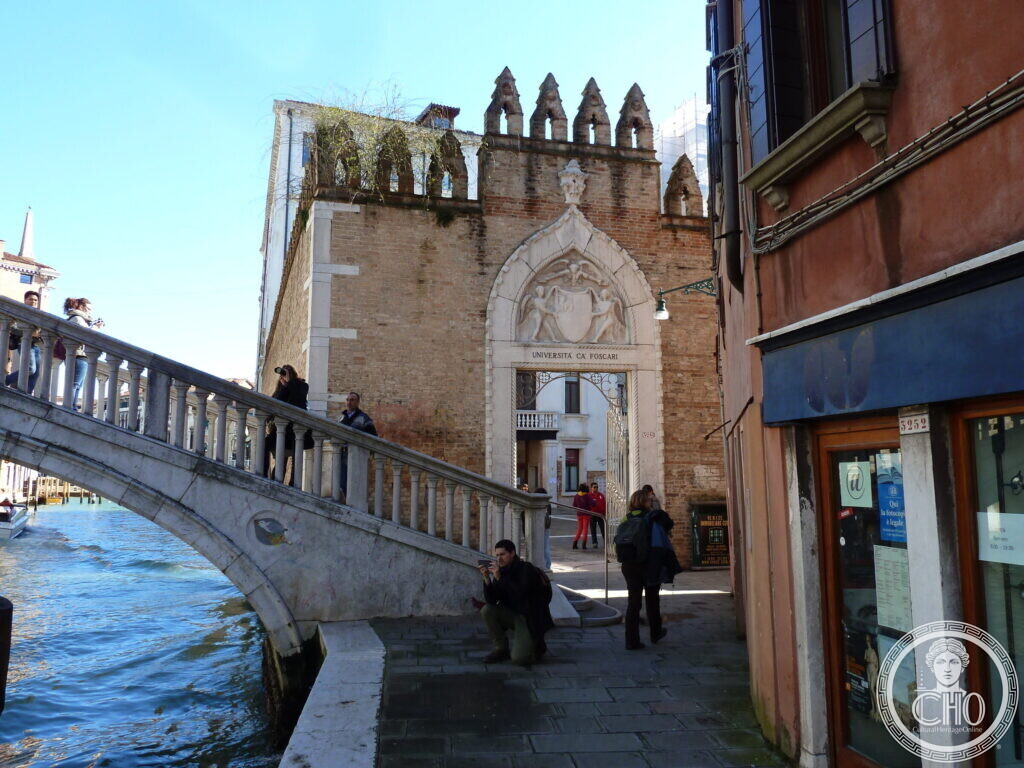 A brick wall topped with Gothic crenellations and a carved stone portal inscribed Universita Ca Foscari, a stone bridge with people leading to it