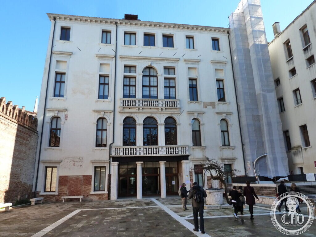 A white palace with arched windows and balconies facing a paved courtyard, a crenellated brick wall on the left and students walking toward the entrance