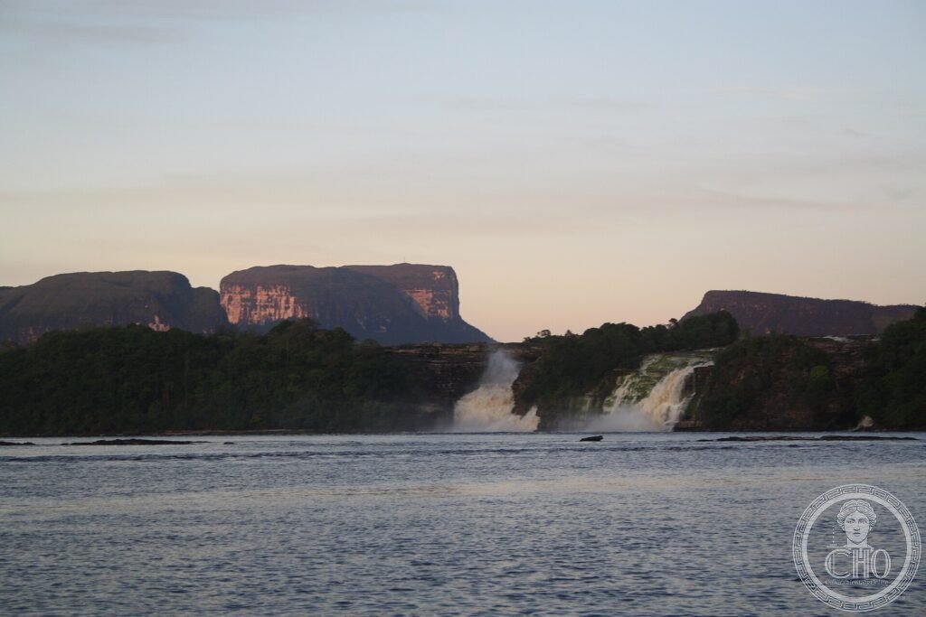 Flat-topped tepui mountains rising at dusk behind waterfalls and a lagoon in Canaima National Park