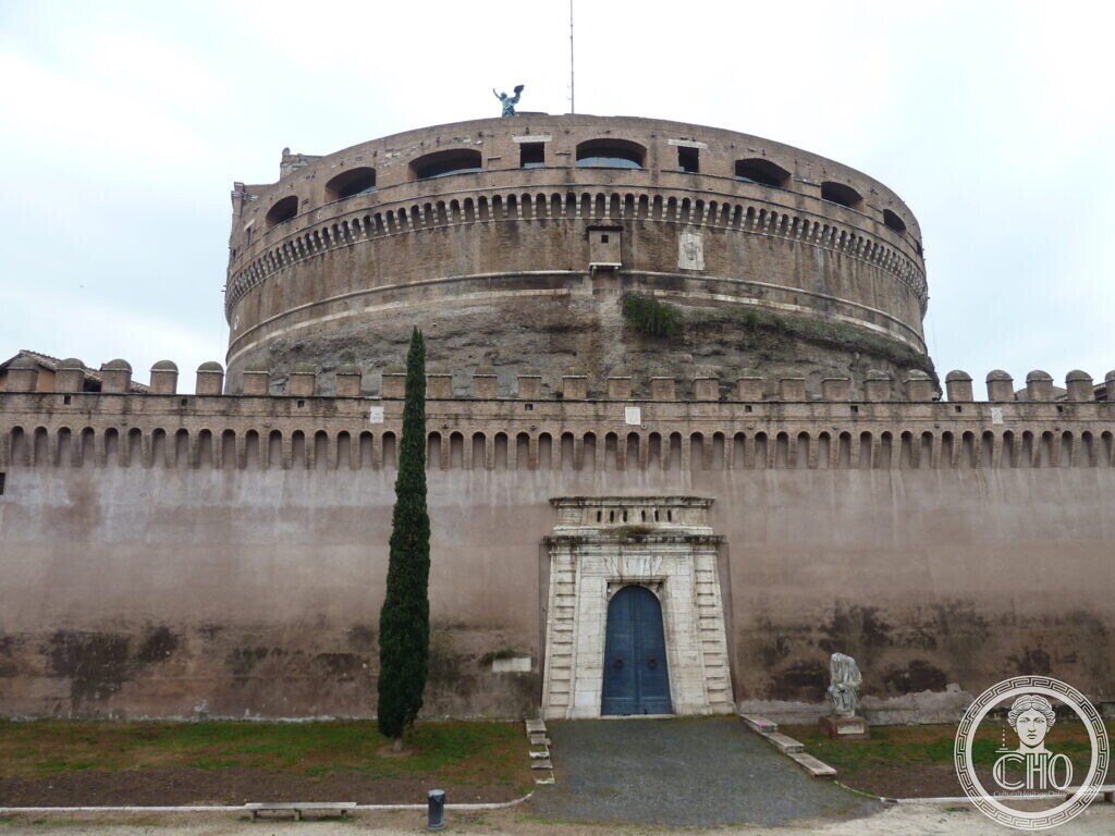 The massive cylindrical core of the castle above a lower crenellated wall with a small arched door and a cypress