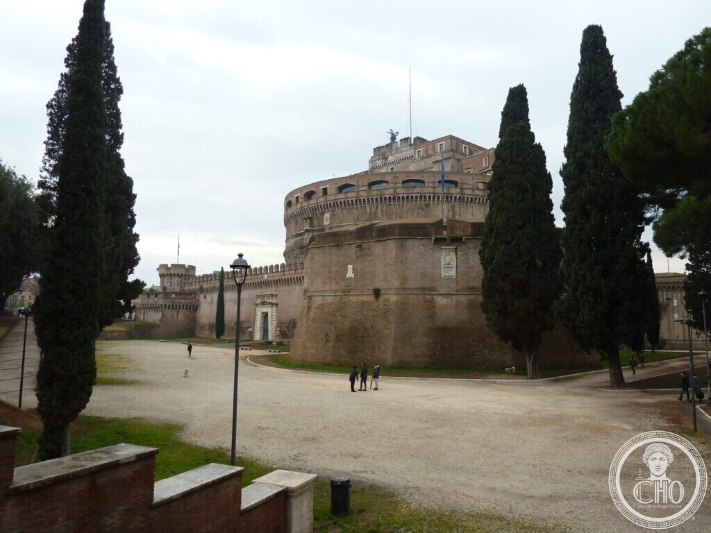 The drum of the castle seen from a gravel path lined with tall cypresses, a small bronze angel on top