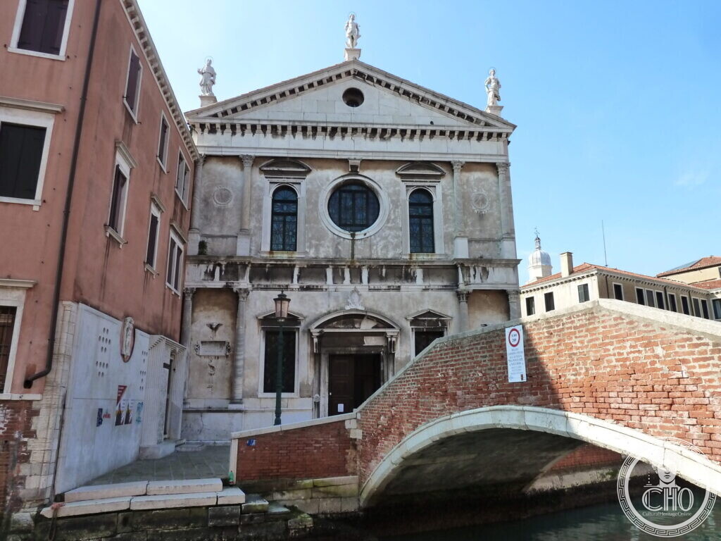 The classical white stone facade of the church of San Sebastiano with statues on the pediment, a red brick bridge over the canal in front