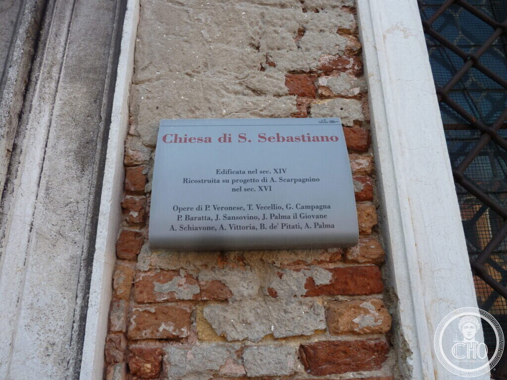 A grey metal plaque on a brick wall headed Chiesa di S. Sebastiano, listing its building dates and the artists whose works it holds