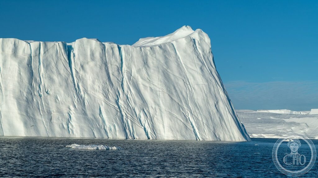 A vast white iceberg with a sheer cliff face floating in the dark water of the Ilulissat Icefjord