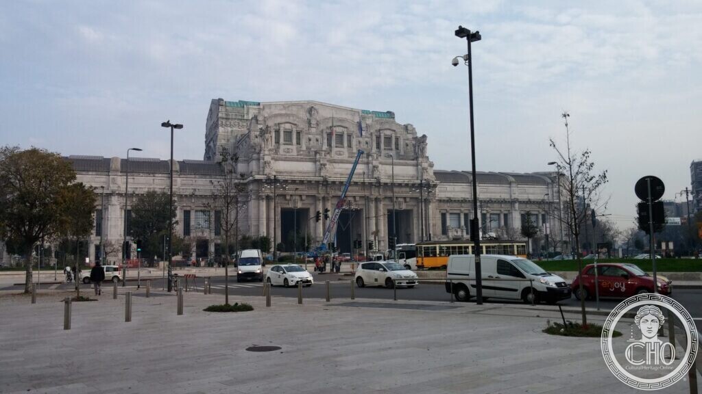 The long stone front of Milano Centrale station with its central block and sculpted attic, seen across the square, cars and a yellow tram in front