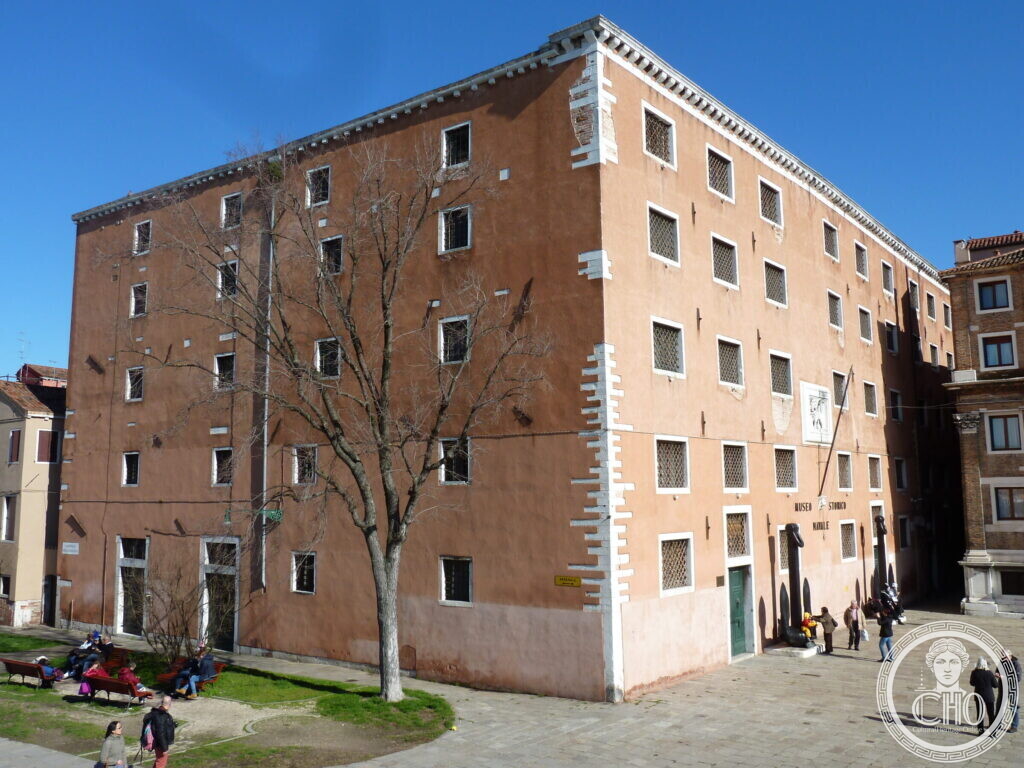 A tall plain pink-plastered building with rows of grated windows and white stone quoins, trees and people in the square in front