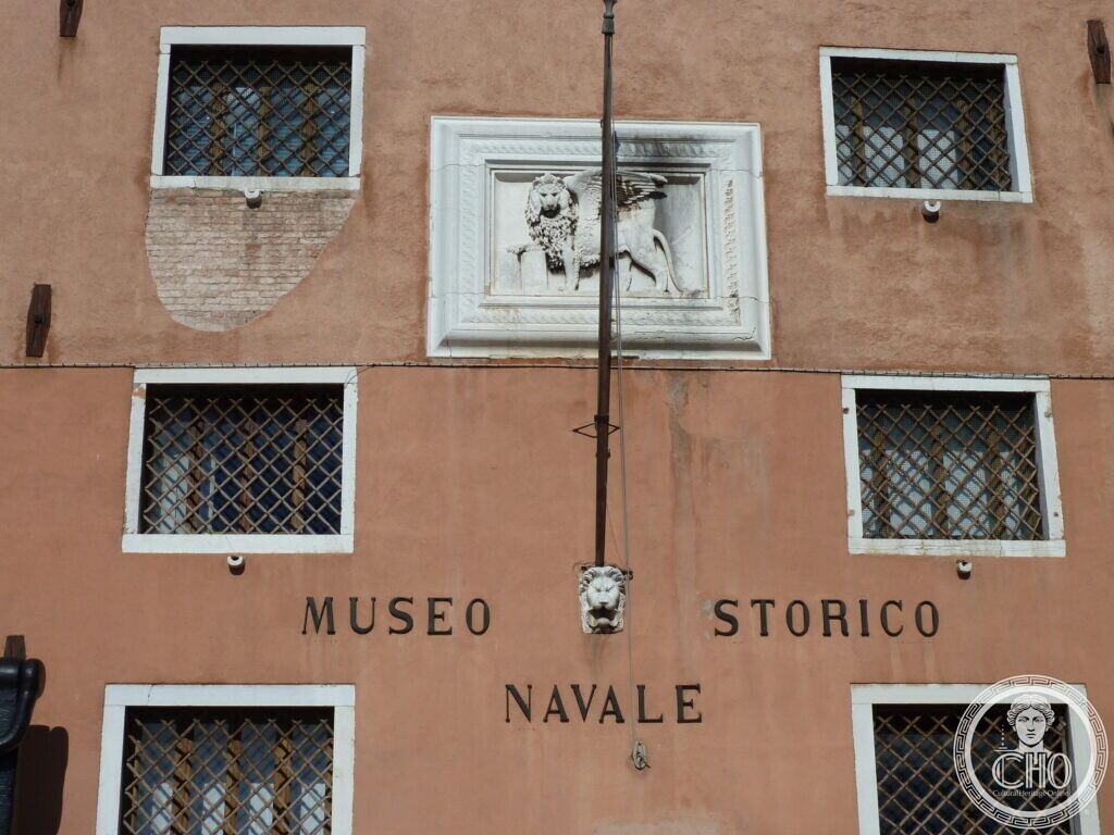 A carved stone panel of the winged lion of Saint Mark on a pink facade, above the words Museo Storico Navale between grated windows