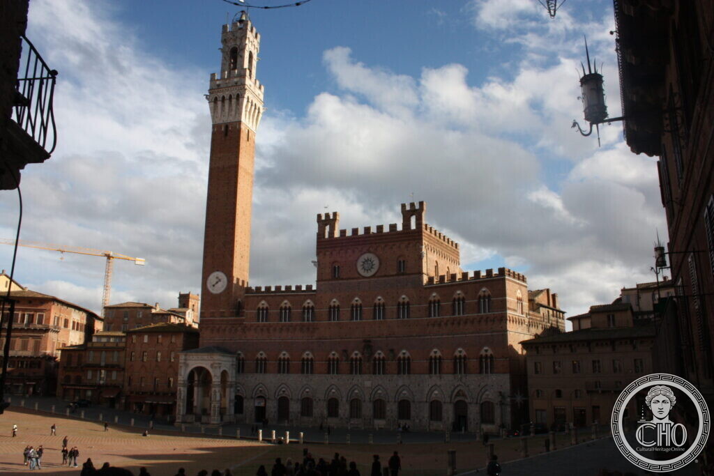 Gothic brick town hall with a crenellated top and a very tall slender tower, seen across the shell-shaped square under a cloudy sky