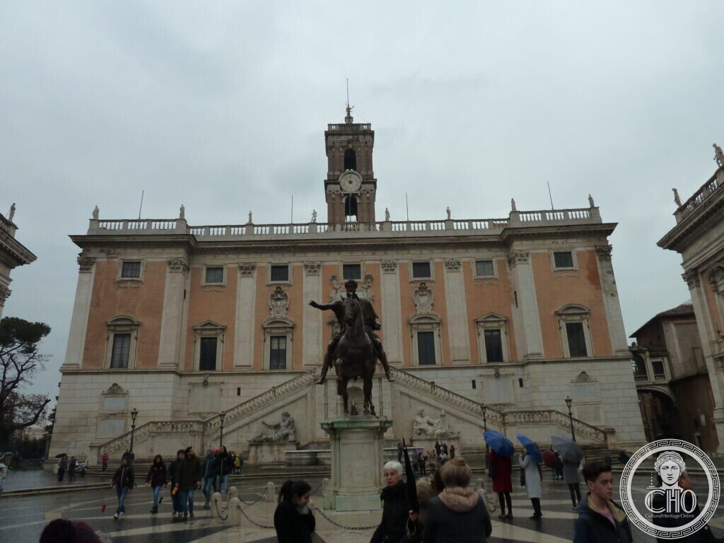 Renaissance palace with a central tower, a double ramp of stairs and a bronze equestrian statue in front, people crossing the square on a grey day