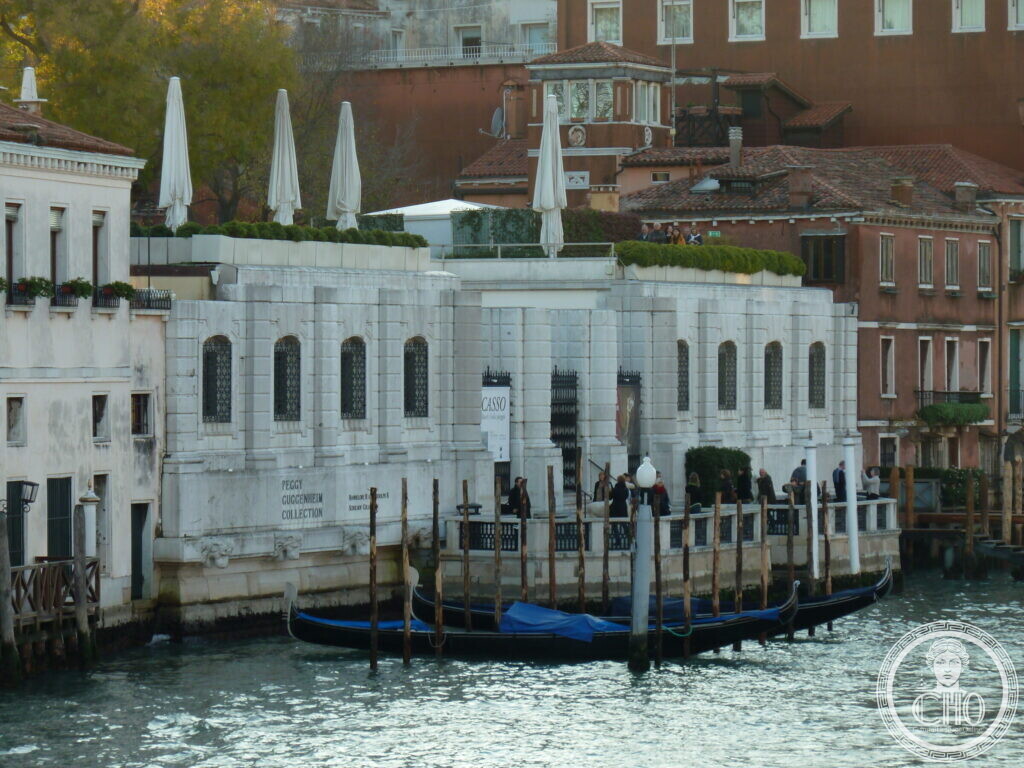 The long low white facade of Palazzo Venier dei Leoni seen across the Grand Canal, with umbrellas on the roof terrace and gondolas in front