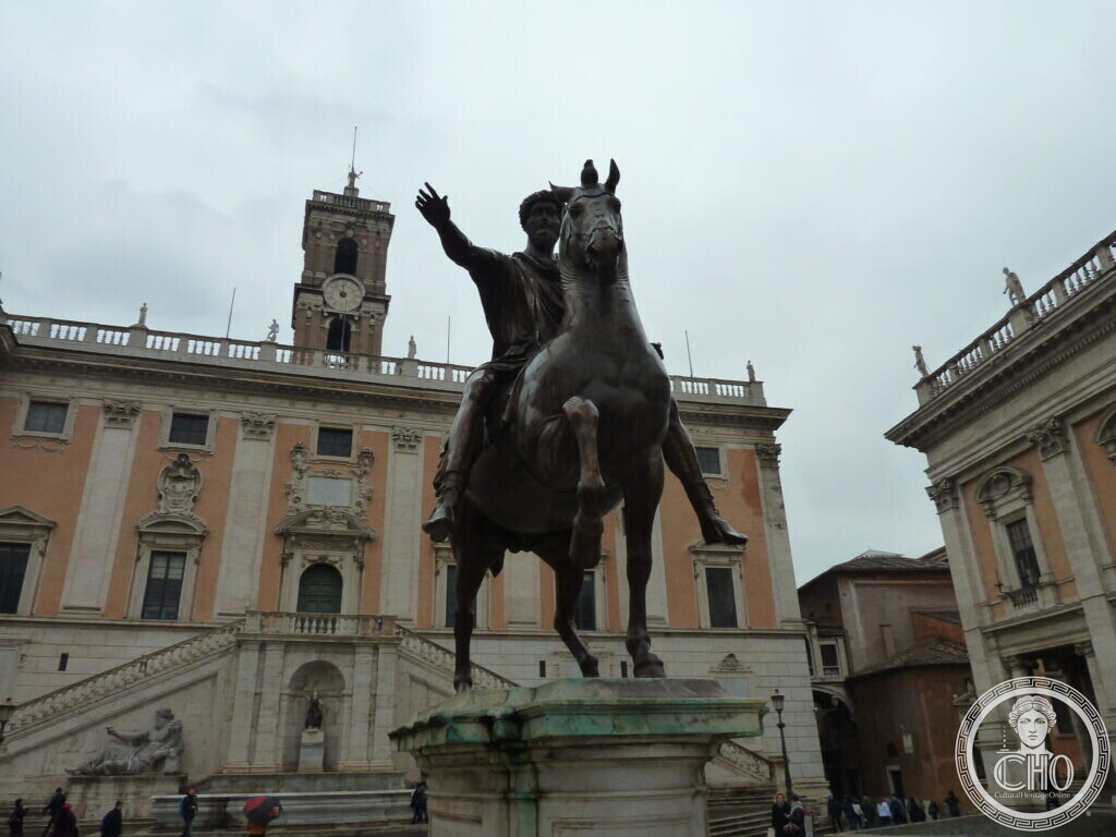 Bronze equestrian statue of an emperor with his arm raised, on a tall marble base, in front of a palace with a clock tower