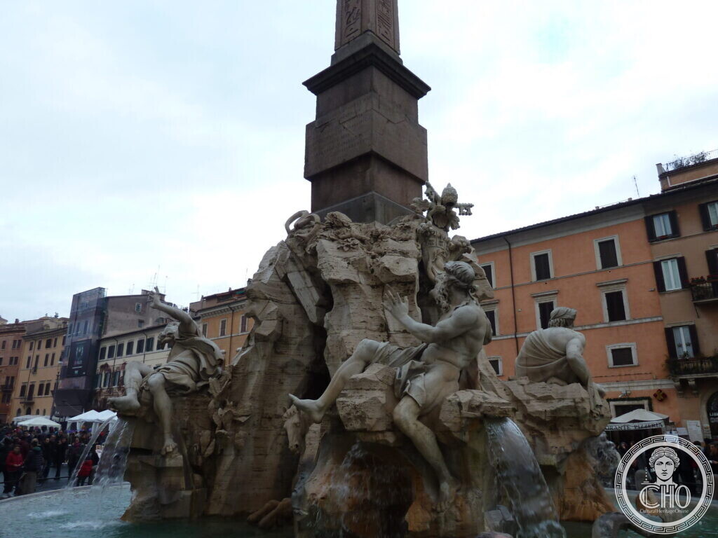 The Four Rivers fountain seen from the side with the obelisk rising above the river gods, house fronts and market stalls behind