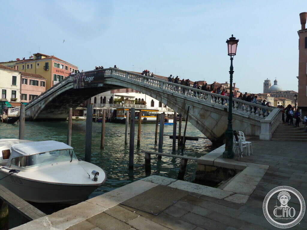 The white stone arch of the Ponte degli Scalzi over the Grand Canal, crowded with pedestrians, a lamp post and moored boat in the foreground