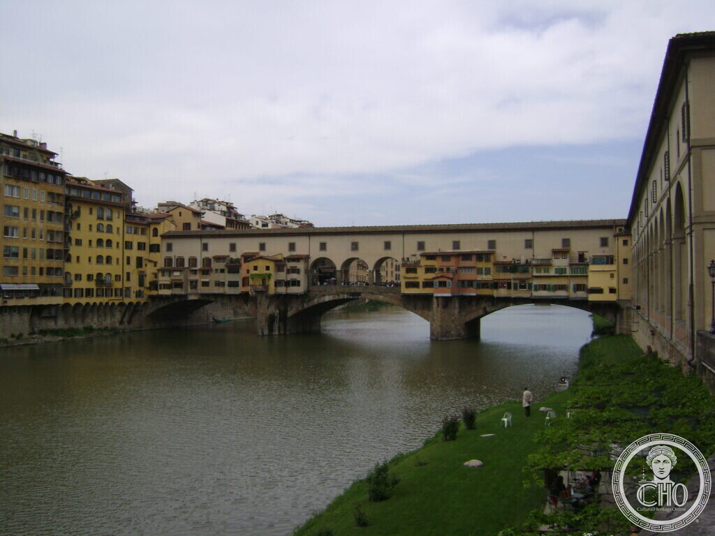 A low three-arched bridge over a green river, lined with shops of different heights and colours, an arcaded building running along the right bank