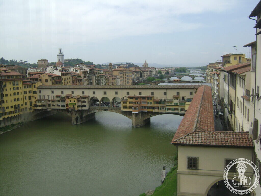 An elevated view of a bridge lined with houses, the Arno curving away towards further bridges and the hills of Florence, a tiled roof in the foreground