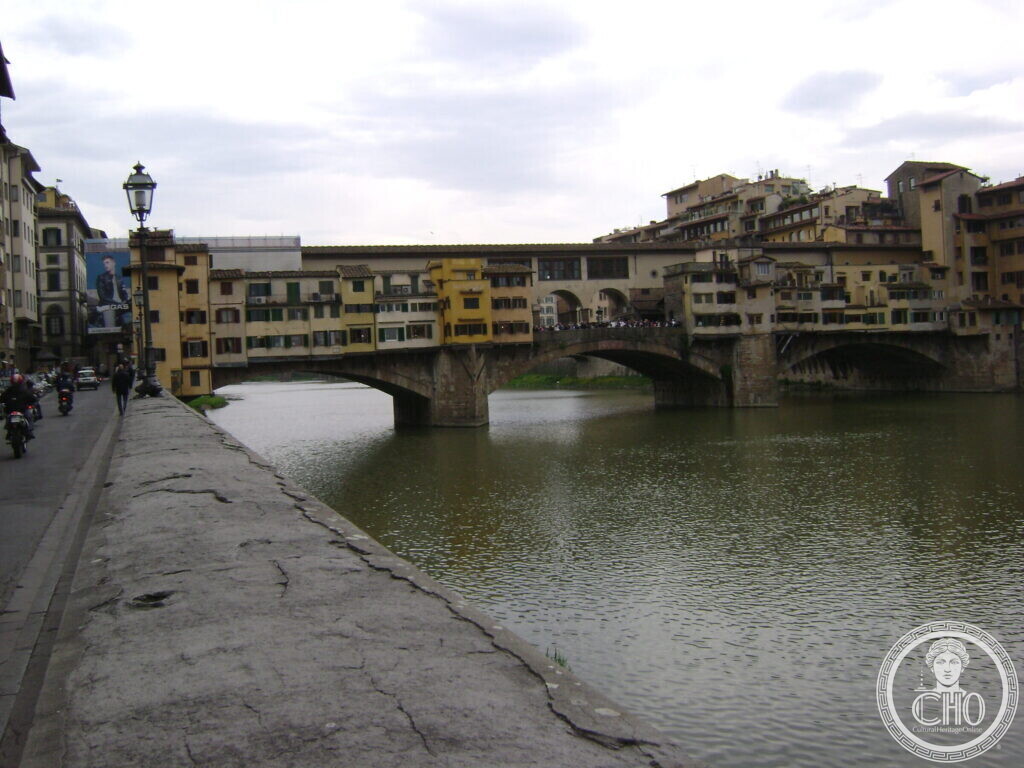 A stone bridge seen from a riverside street, with yellow and ochre shop fronts projecting over the water on wooden brackets