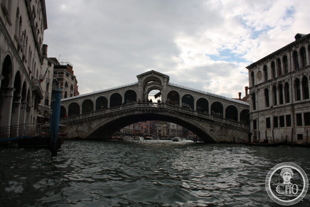 The Rialto Bridge seen from the water at a low angle, its central arch and arcades in silhouette against a bright sky