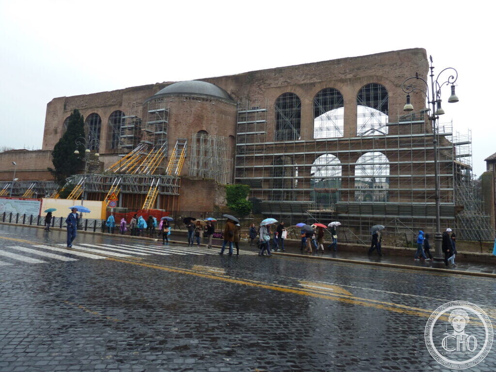 Three enormous brick coffered vaults of a Roman basilica seen from the street, partly covered by scaffolding, people with umbrellas on the pavement