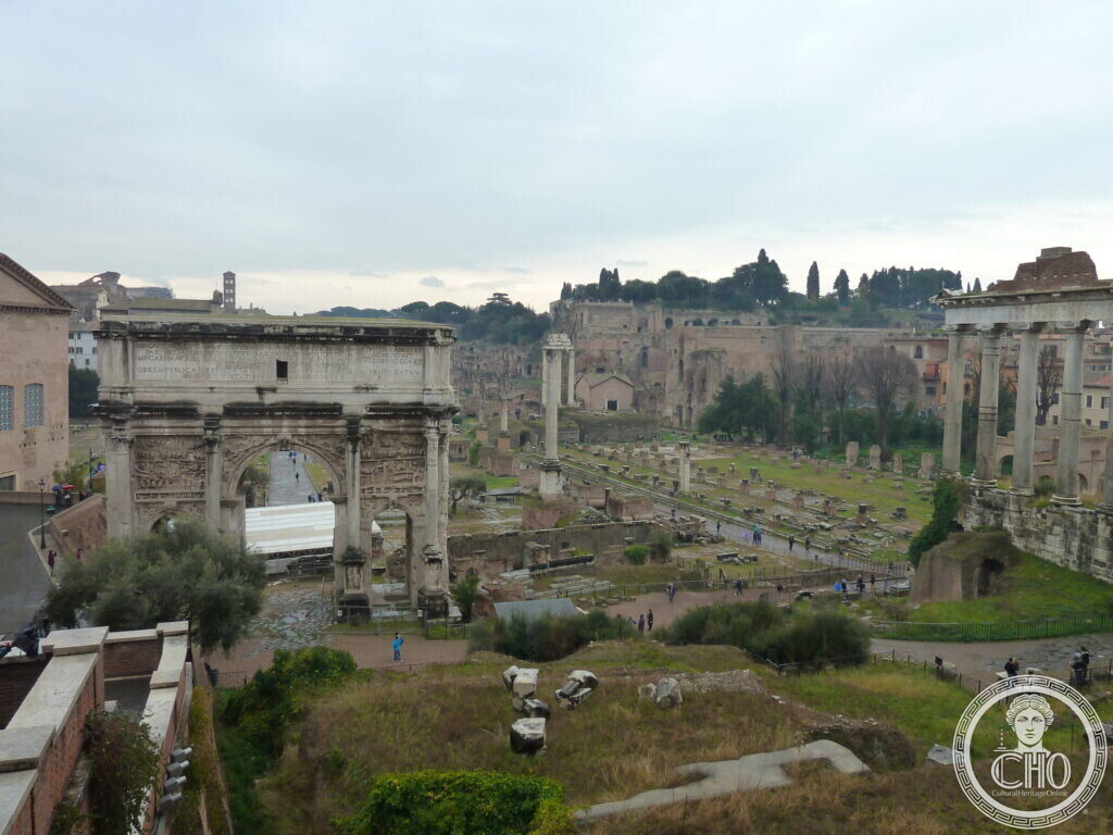 Wide view of the Roman Forum from above with the Arch of Septimius Severus on the left, scattered columns and ruins, and the Palatine hill in the distance