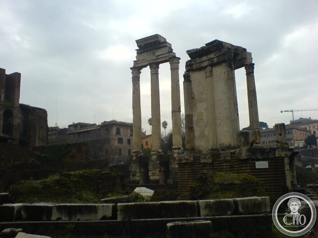 Three fluted Corinthian columns carrying a piece of entablature, with a smaller reconstructed temple front to the right, under a grey sky
