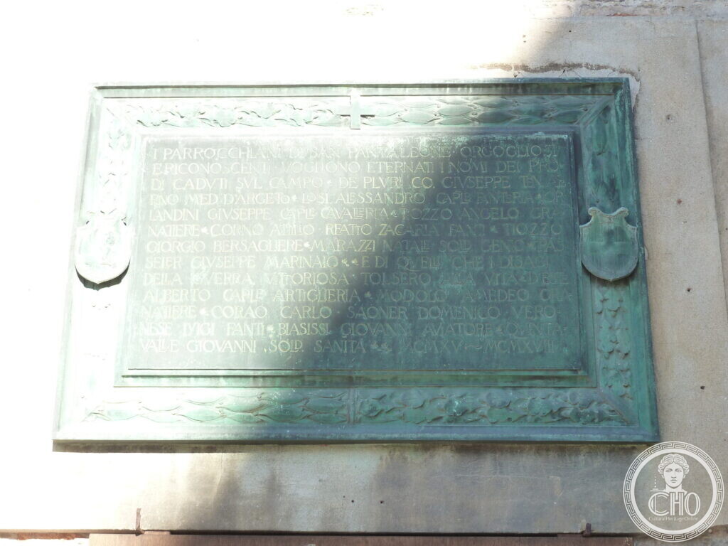 A green bronze plaque with a raised border of leaves and two shields, listing names of fallen soldiers, dated MCMXV to MCMXVIII