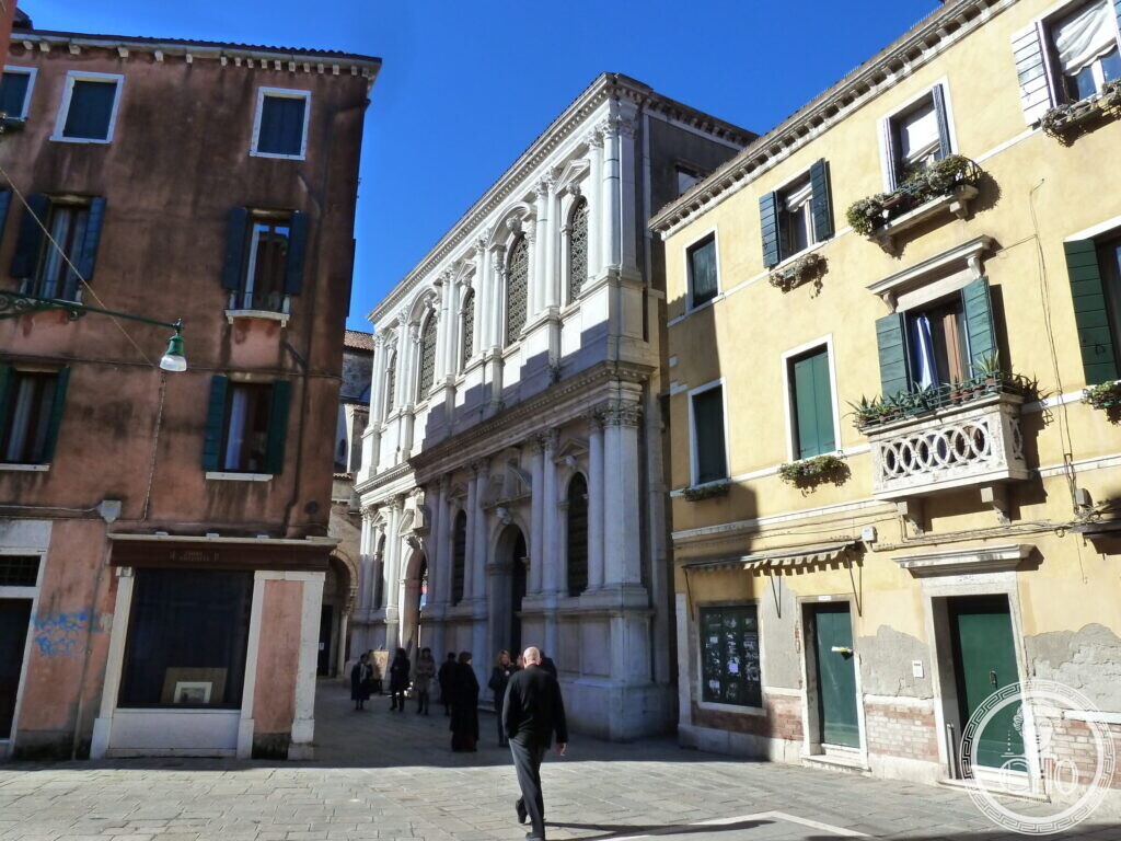 The white marble facade of the Scuola Grande dei Carmini between a pink and a yellow house, people walking across the paved square