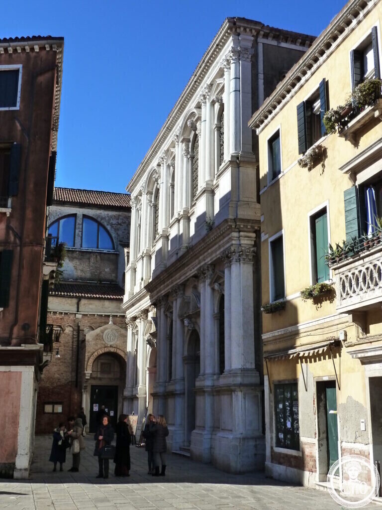 A tall white marble facade with two orders of columns between plain houses, the brick church of the Carmini visible at the end of the square