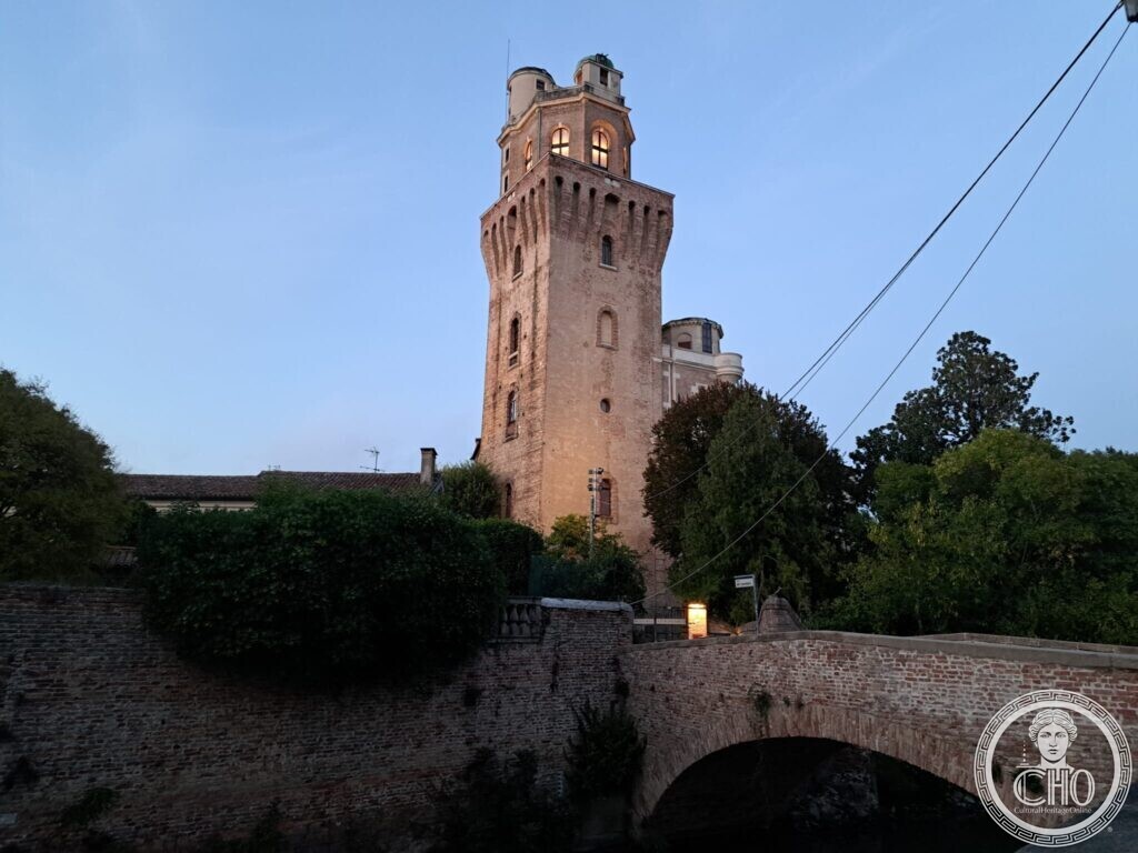 The tower of the Specola at dusk with lit windows, seen from a brick bridge whose parapet fills the foreground