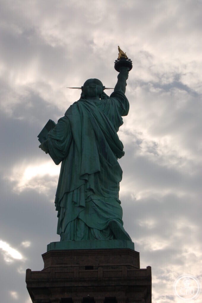 The Statue of Liberty seen from below in silhouette against a bright cloudy sky, the torch raised