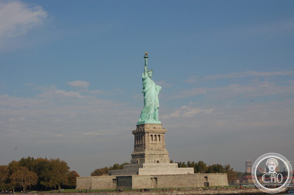 The Statue of Liberty on its granite pedestal and star-shaped fort, seen from the water with a shoreline behind
