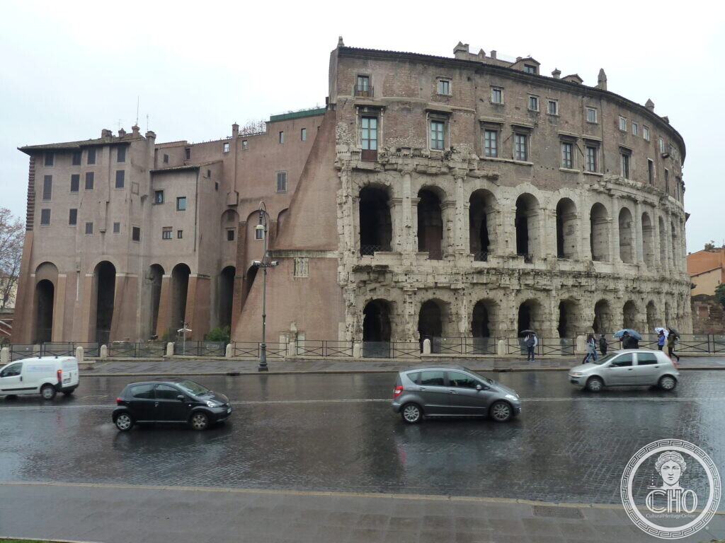 Two tiers of Roman arches with a later palace on top, cars passing on the wet road in front