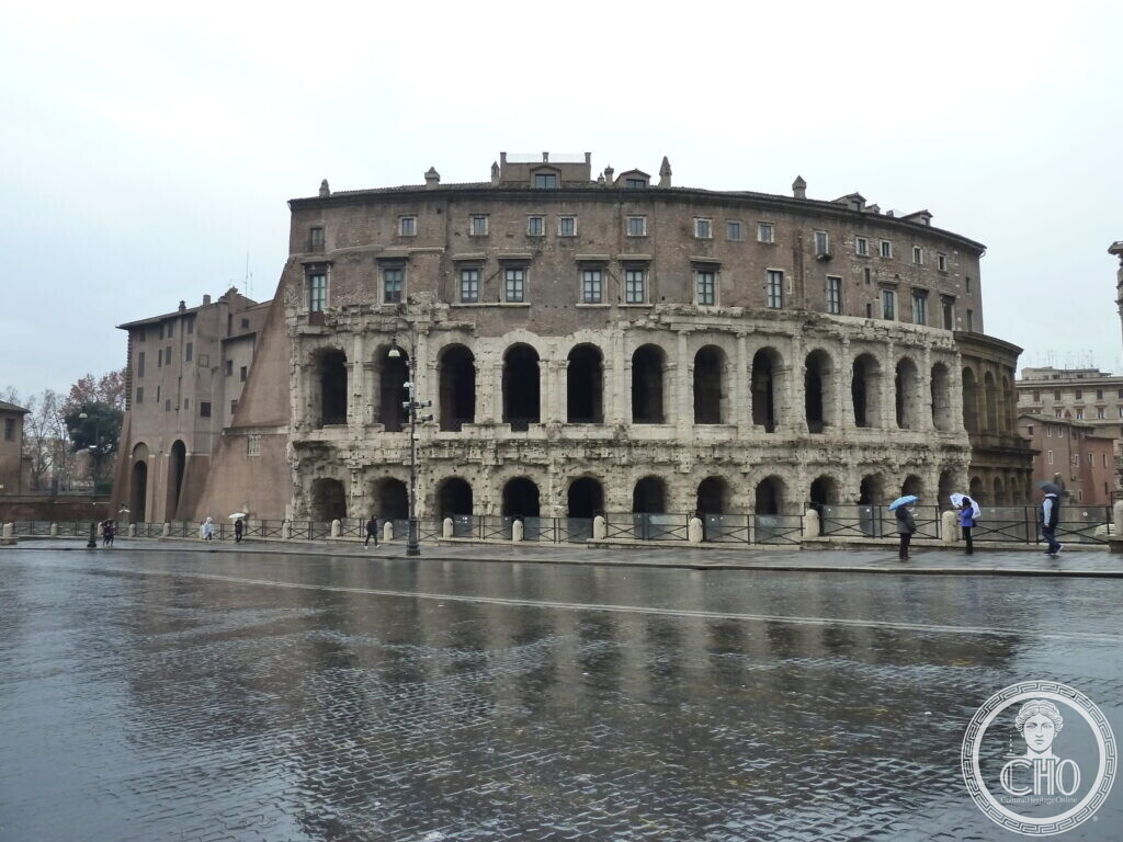 Curved ancient theatre front with two tiers of arches in travertine, a Renaissance palace rising above them, wet street in the foreground