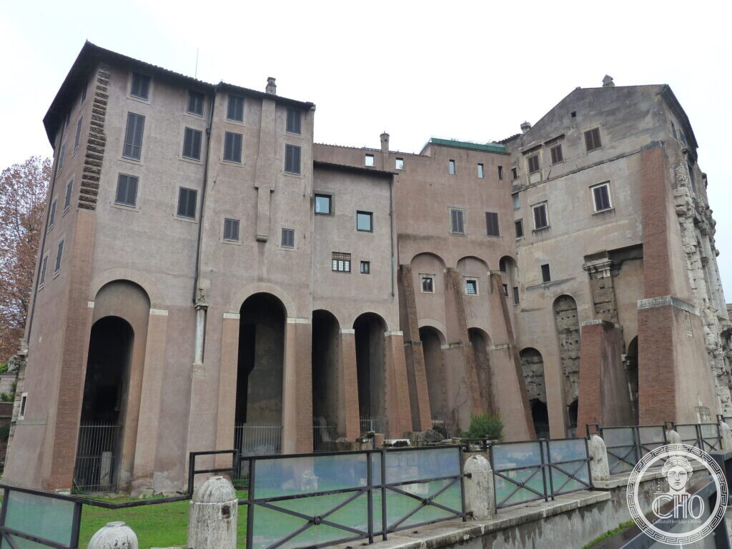 Tall old houses with arched openings built against the theatre, three white columns with entablature standing on the right