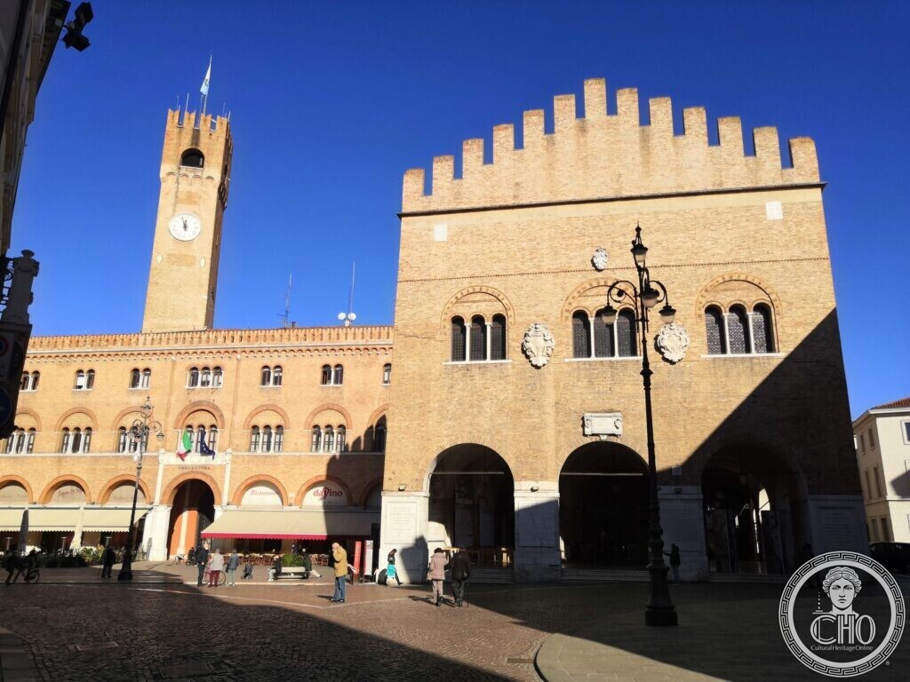 A brick medieval palace with swallowtail battlements and open arches, next to a long arcaded palace and a clock tower, people in the square